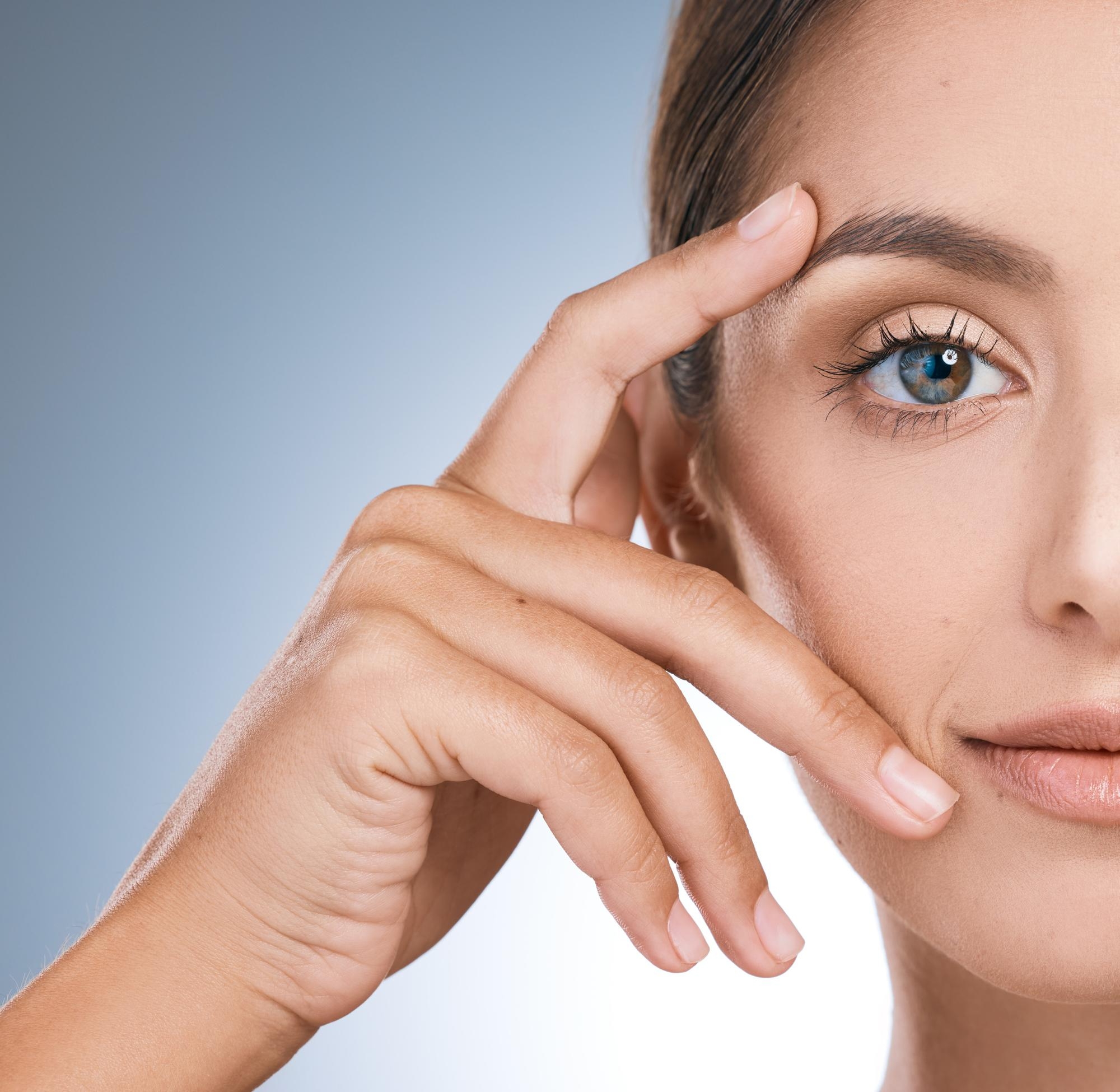 Half-face beauty portrait of a woman framing her eye and cheek with her hand against a soft blue background