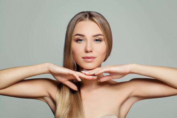 Beauty portrait of a blonde woman facing forward with both hands placed under her chin against a light gray background.