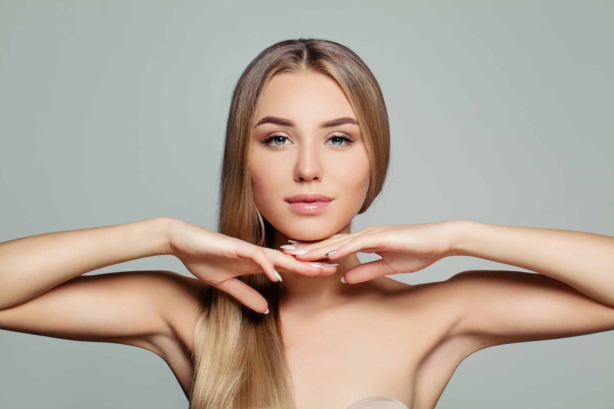 Beauty portrait of a blonde woman facing forward with both hands placed under her chin against a light gray background.