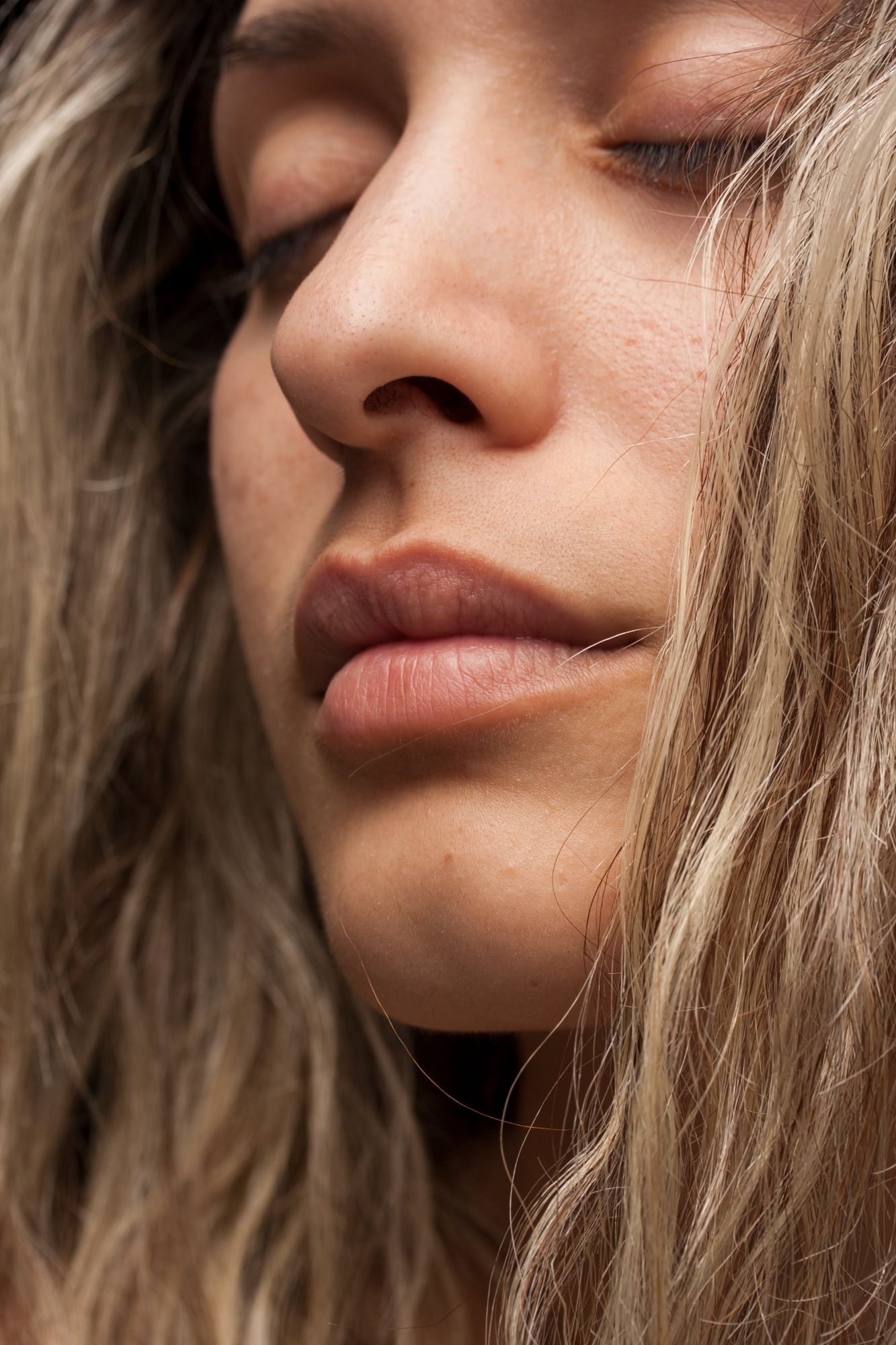 Close-up portrait showing natural skin texture, nose, and lips framed by blonde hair.