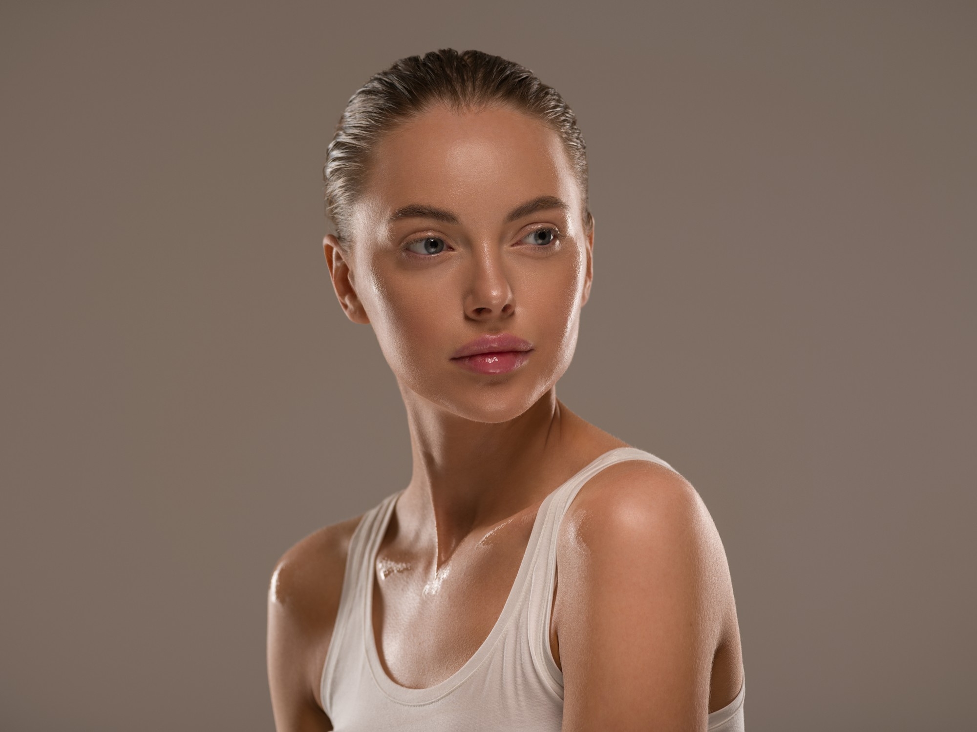 Portrait of a woman with glowing skin wearing a white tank top and looking to the side against a neutral background.
