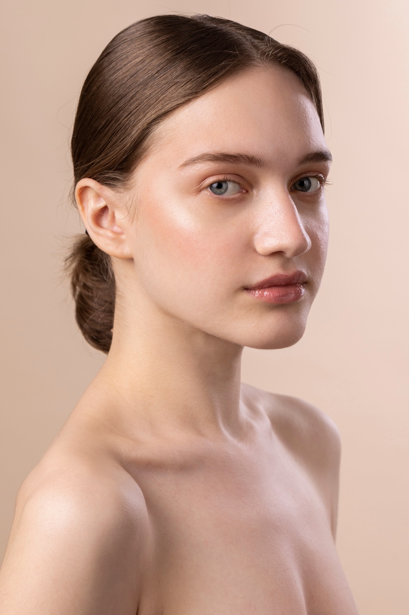 Front portrait of a brunette woman with minimal makeup and bare shoulders against a soft beige background.
