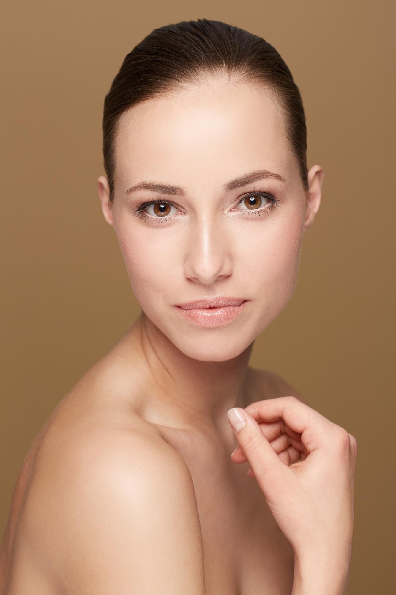 Natural beauty portrait of a woman with pulled-back hair and bare shoulders against a brown background.