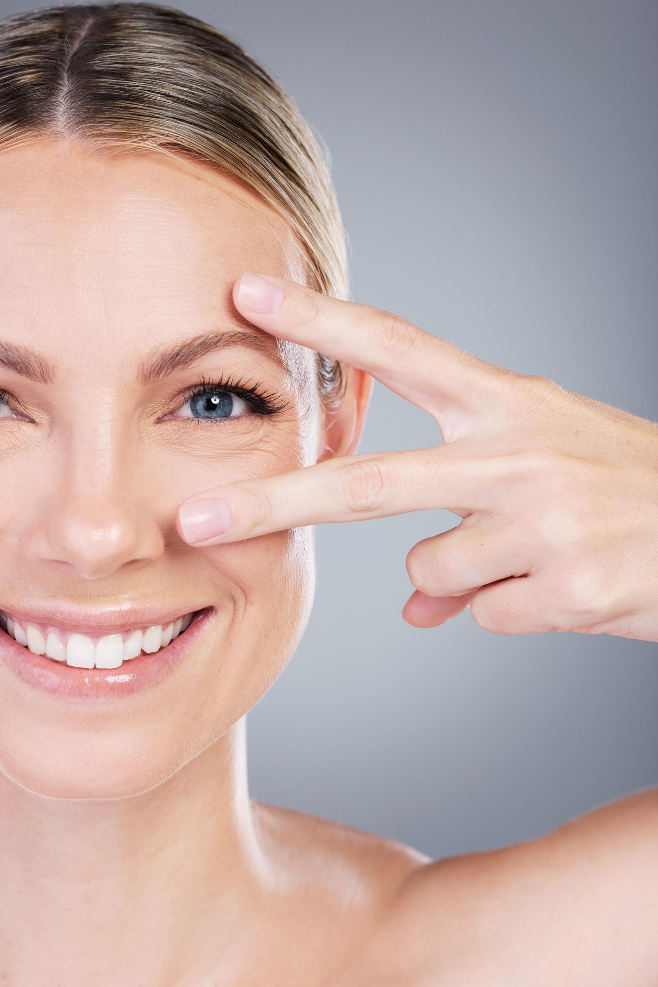 Close-up of a smiling woman framing her eye area with two fingers against a light gray background.
