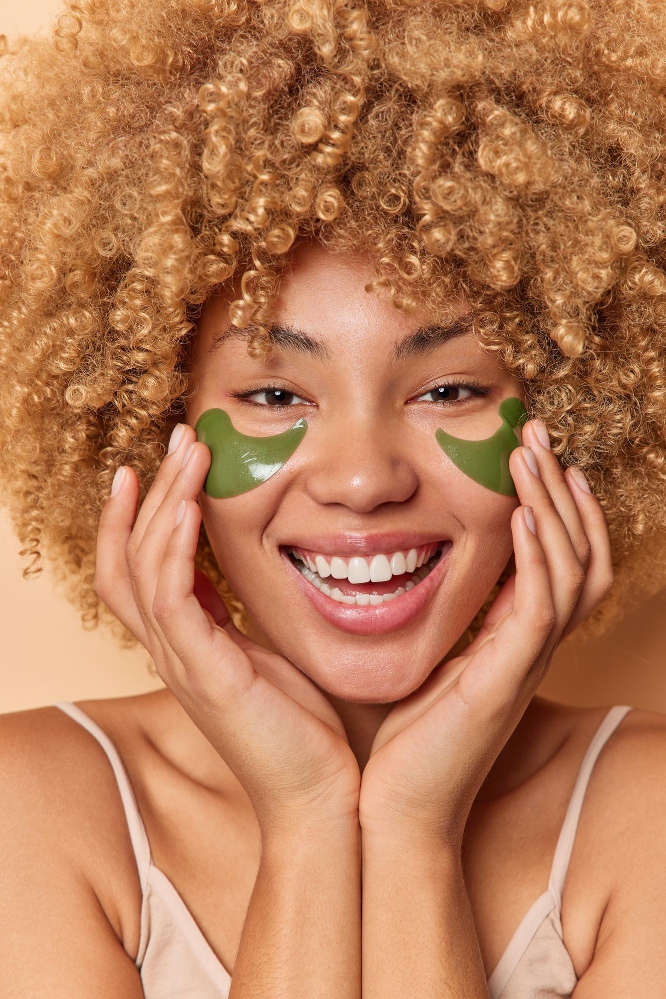 Smiling woman with curly hair applying green under-eye patches in a skincare beauty portrait