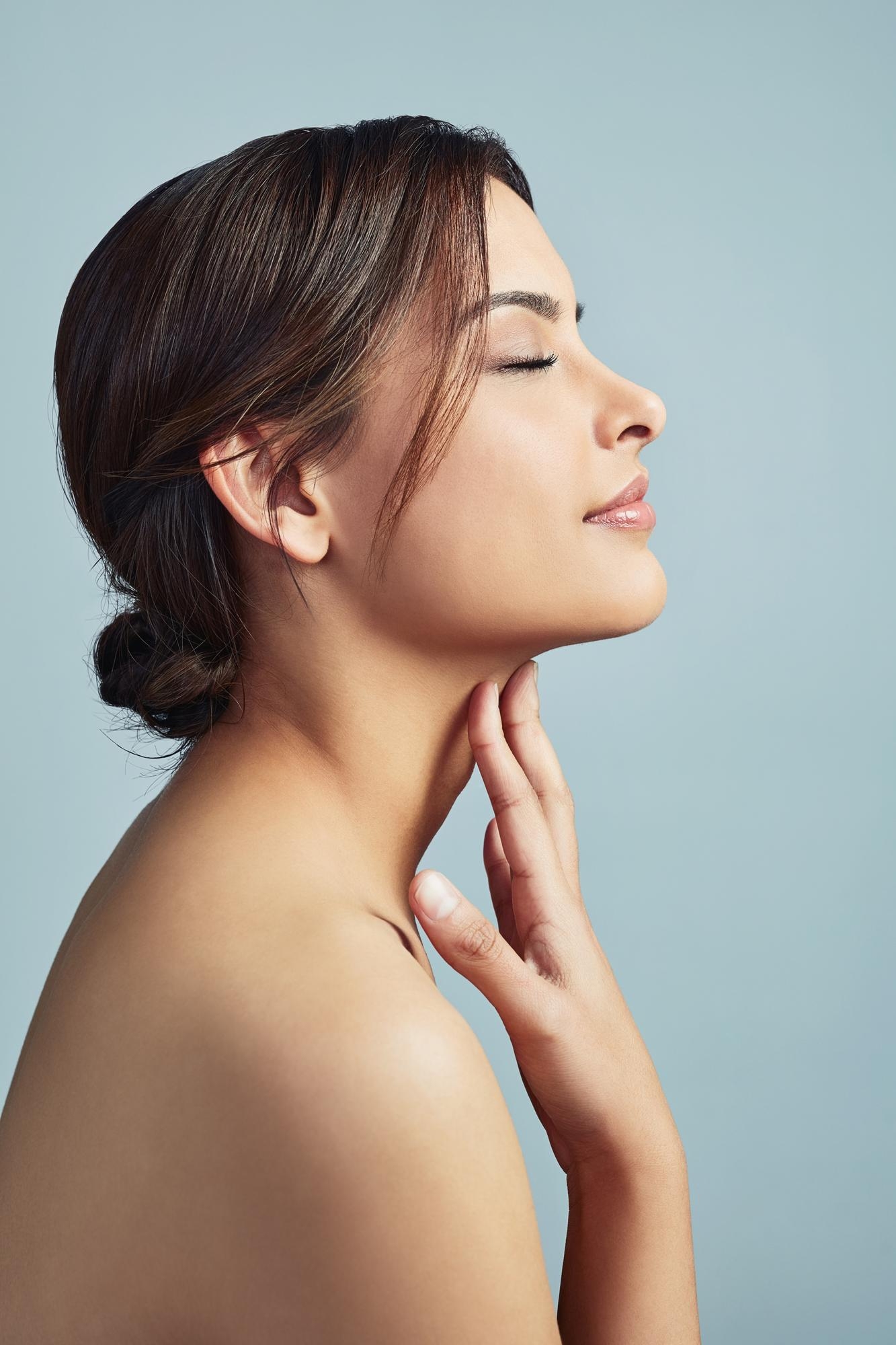 Side profile of a brunette woman with eyes closed resting her hand on her neck against a pale blue background.