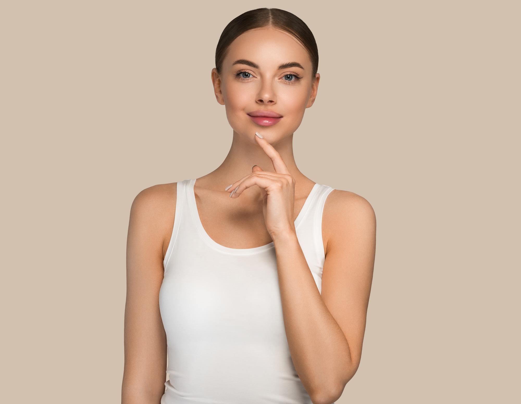 Portrait of a woman in a white tank top gently touching her chin against a beige background.