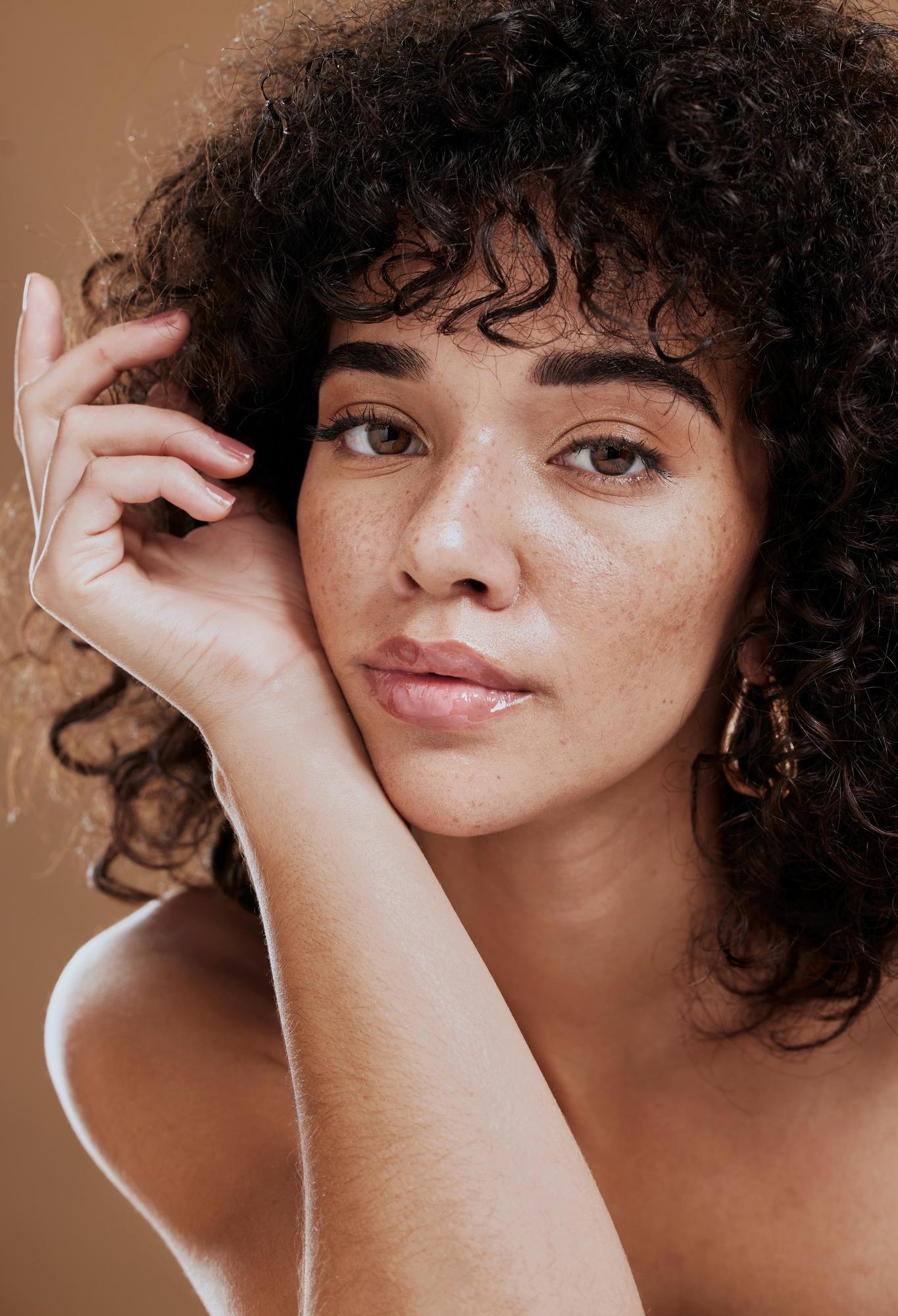Close-up portrait of a woman with curly hair and freckles resting her face against her hand on a warm neutral background.