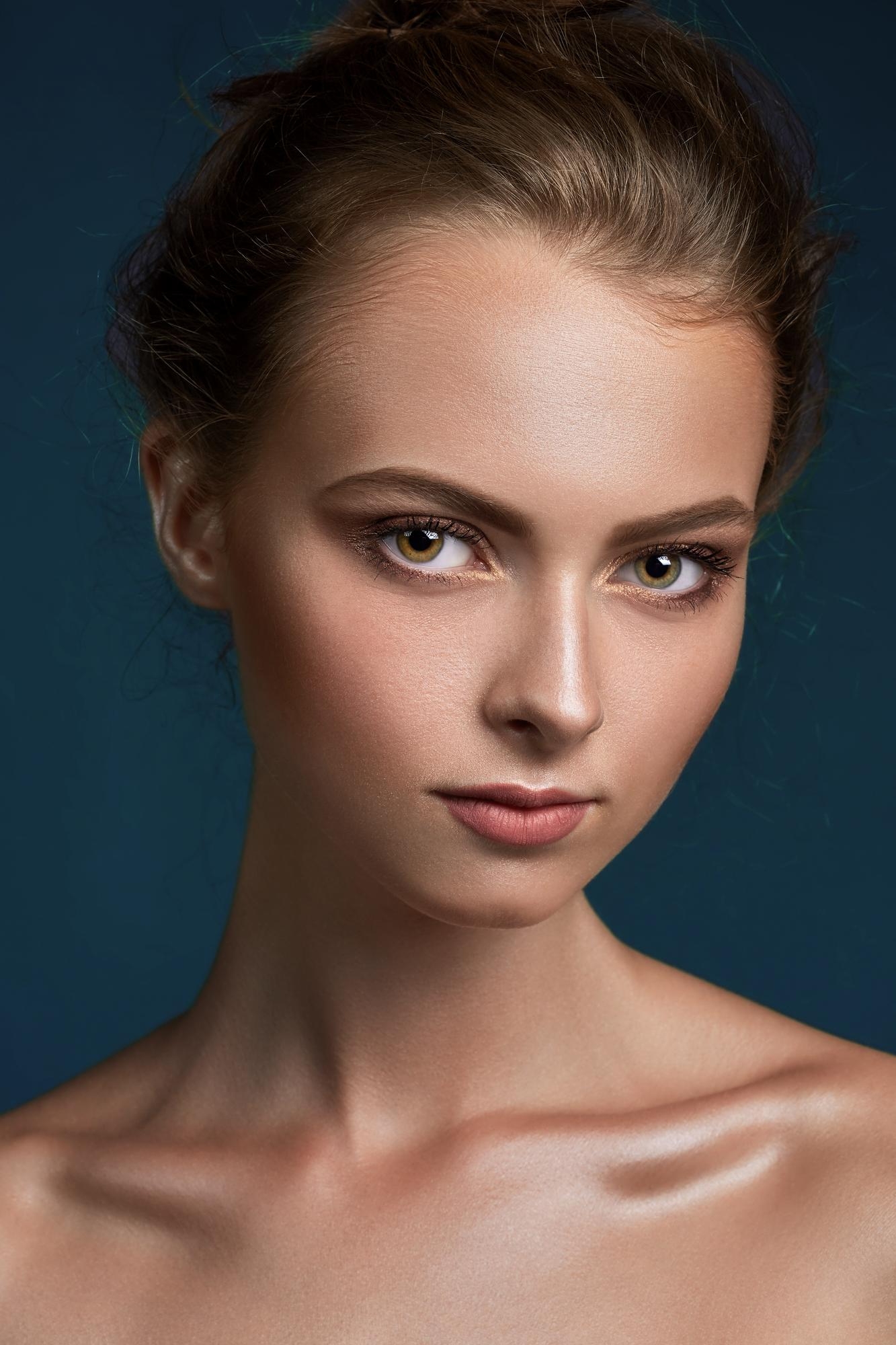 Close-up editorial beauty portrait of a woman with an updo hairstyle, defined eye makeup, and a dark blue background.