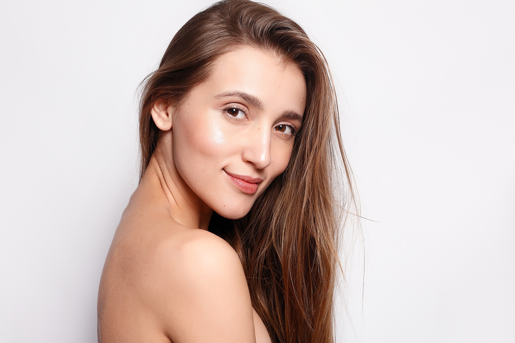 Bright skincare beauty portrait of a woman with long brown hair, glowing skin, and a soft smile against a white background.