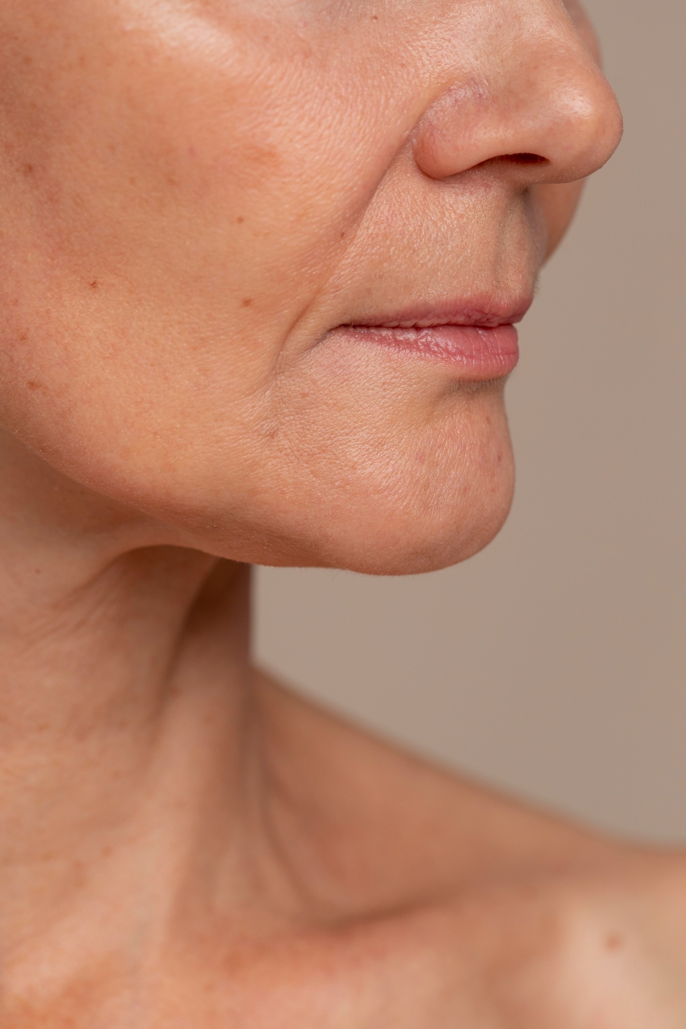Close-up side profile of mature skin showing natural texture, fine lines, lips, and jawline against a neutral background.