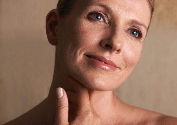 Close-up skincare portrait of a mature woman gently touching her neck, with soft light emphasizing natural skin texture.