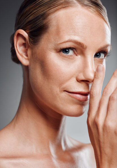Close-up portrait of a woman with slicked-back hair touching her face, showing smooth, glowing skin against a gray studio background.