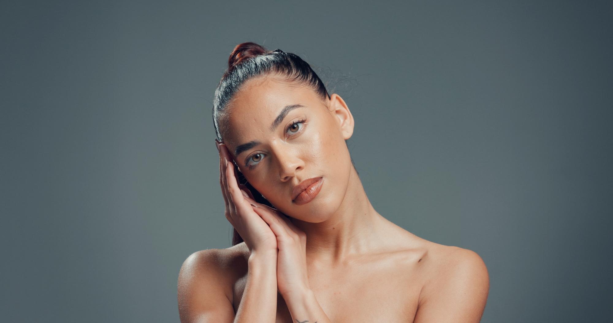 Studio beauty portrait of a woman with glowing skin holding her hands against her cheek against a gray background