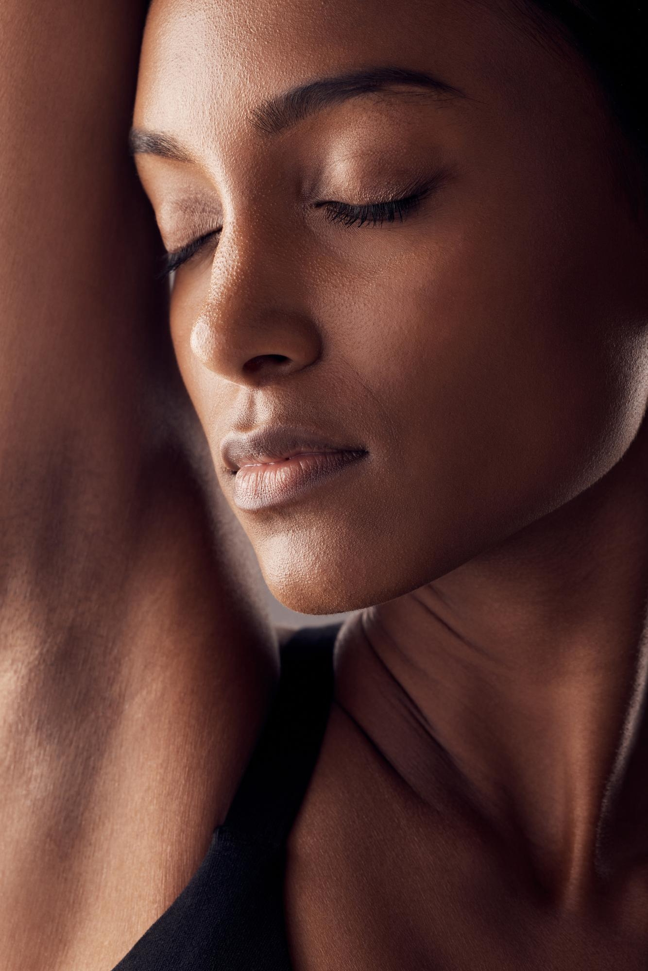 Dramatic close-up portrait of a woman with closed eyes and glowing skin, with her arm raised beside her face.