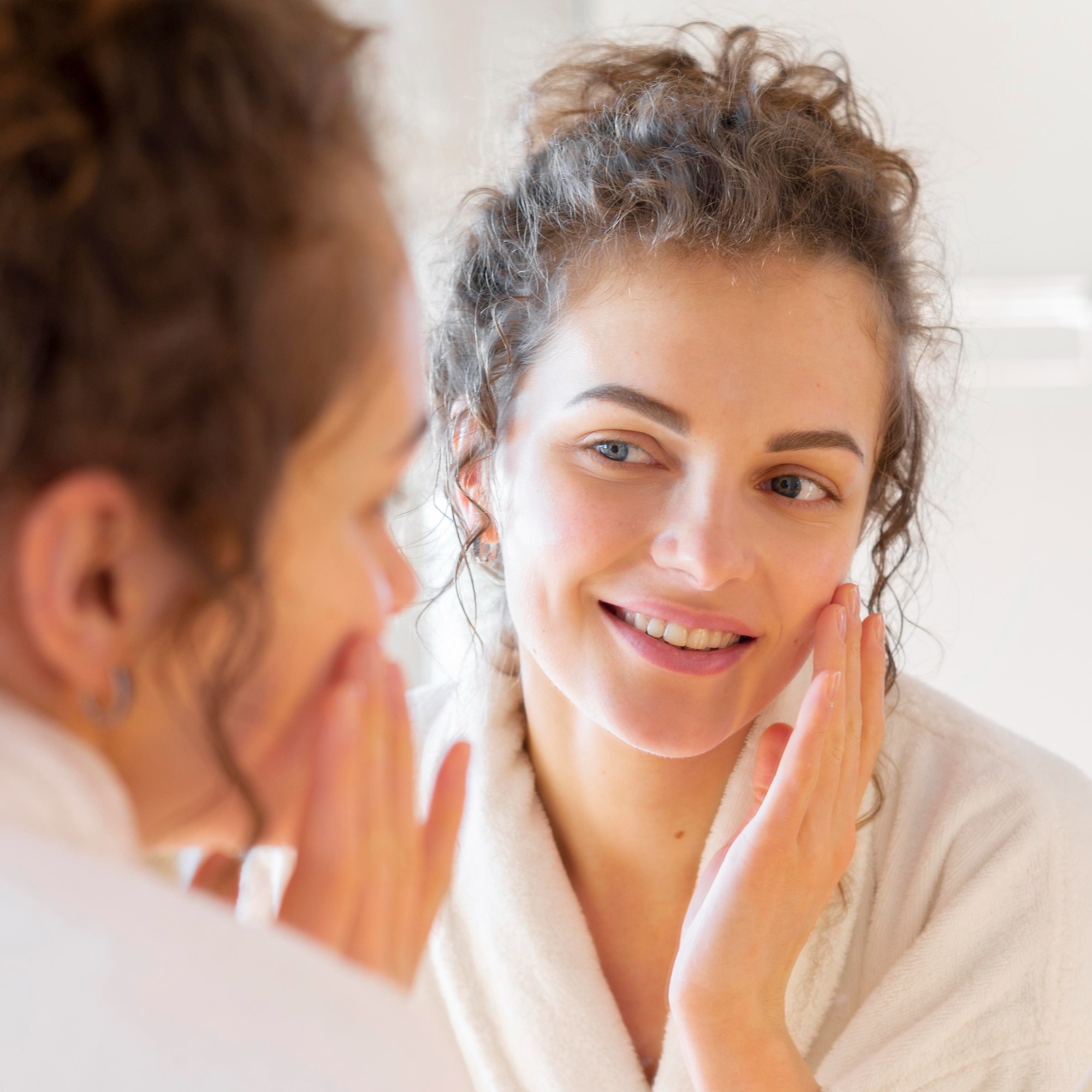 Woman in a white robe smiling at her reflection while gently touching her cheek during a skincare routine.