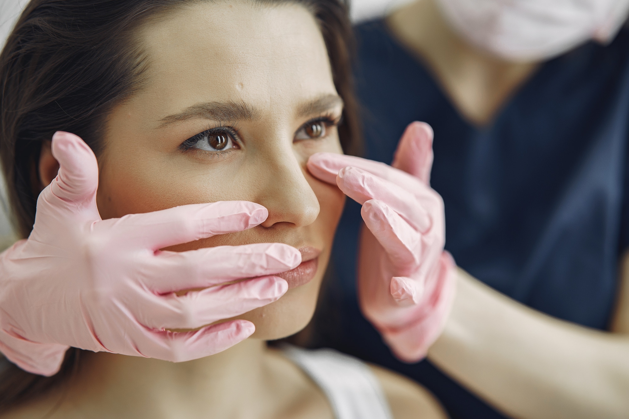 Aesthetician in pink gloves examining a female patient's face during a skin consultation.