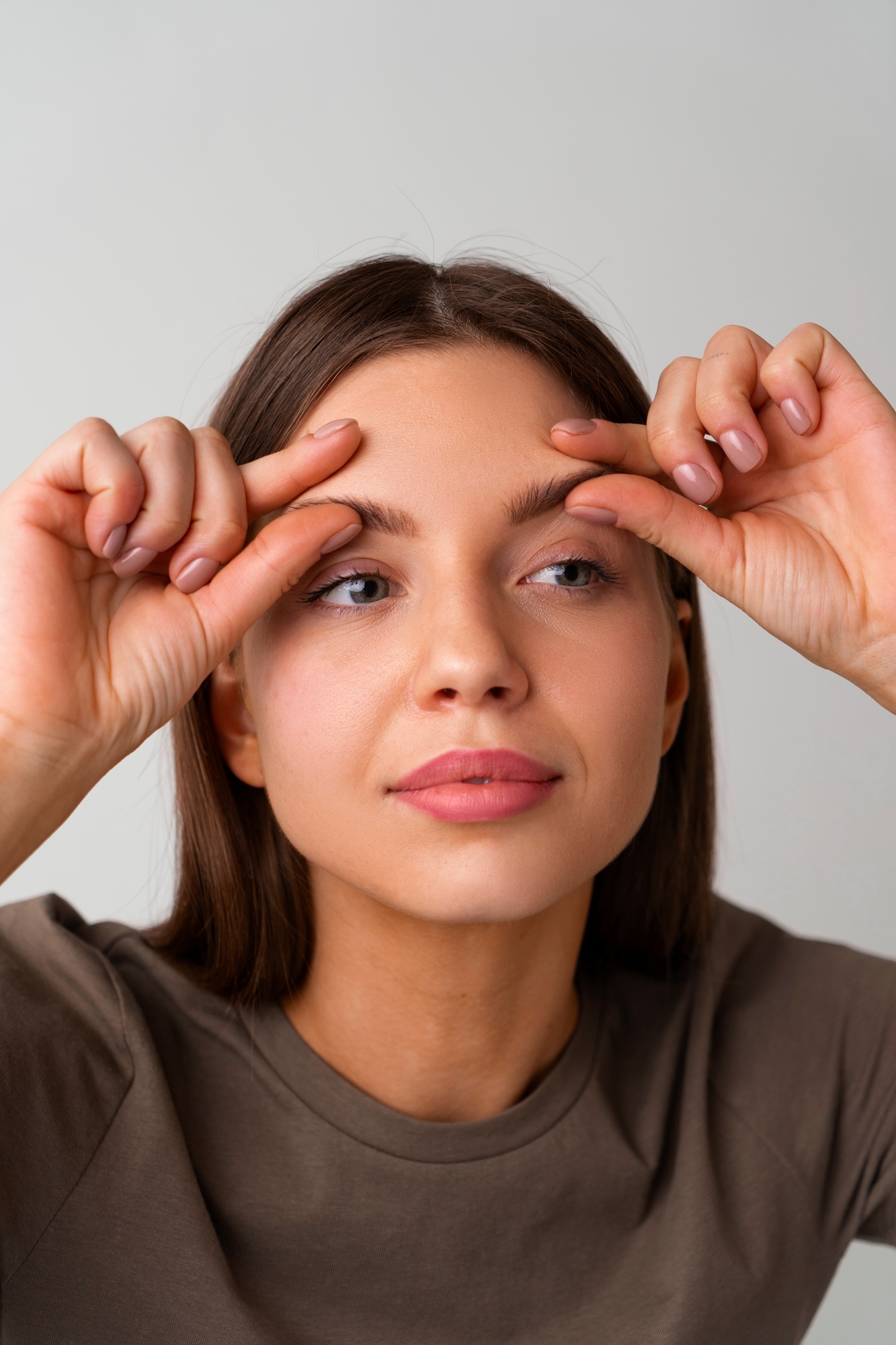 Woman touching her eyebrows with both hands against a light gray background.