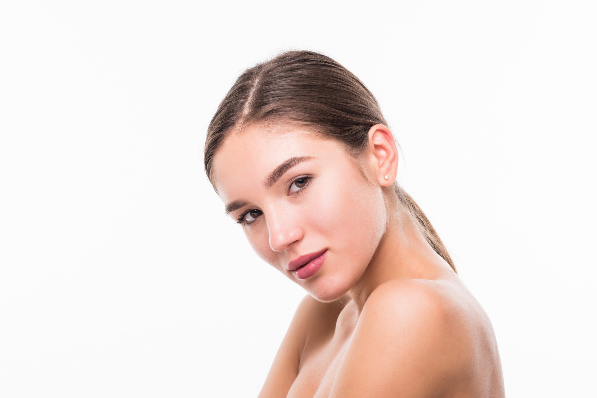 Angled beauty portrait of a young woman with a ponytail and clear skin against a white background.
