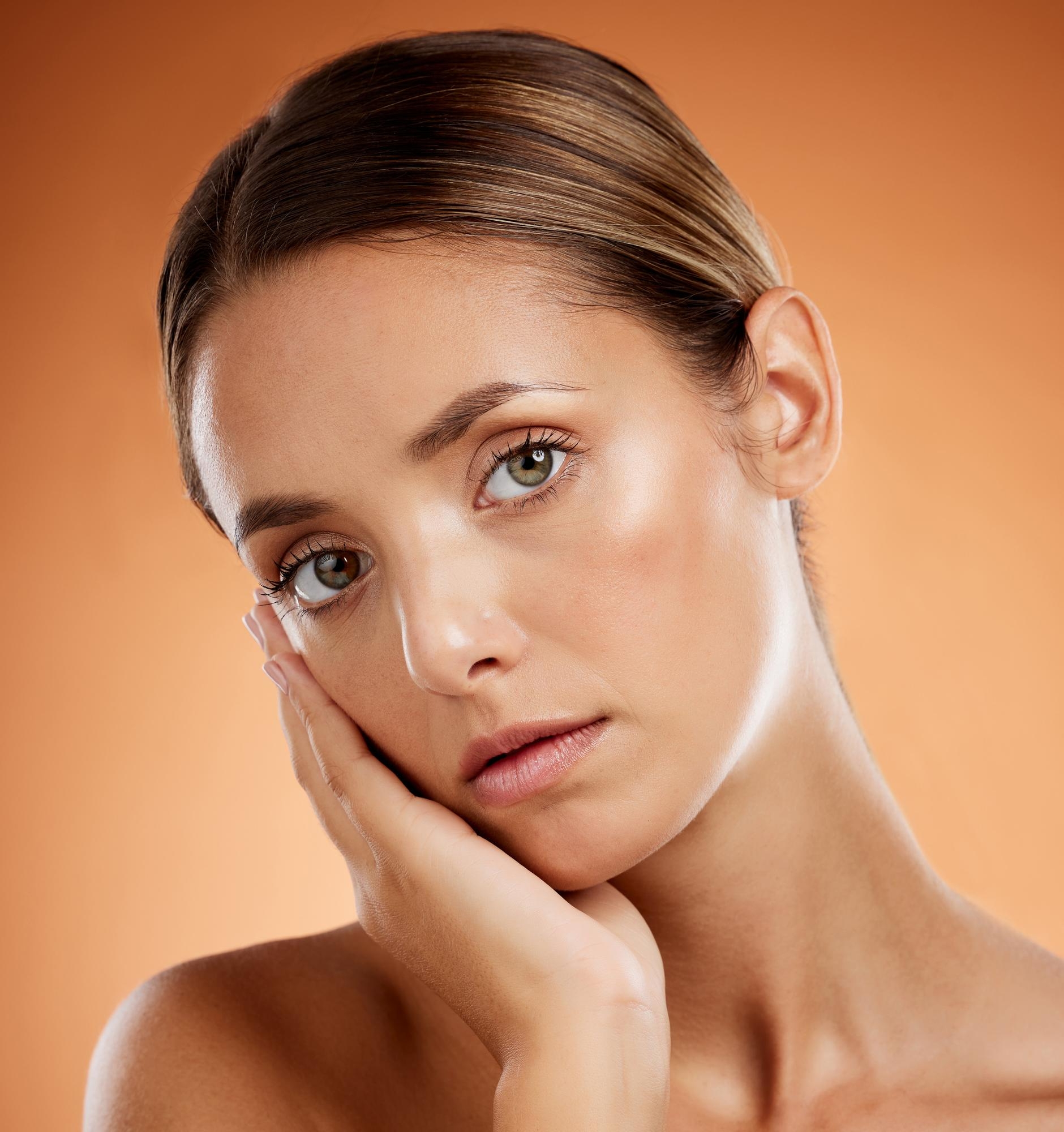 Studio beauty portrait of a woman with glowing skin resting her hand on her face against an orange background