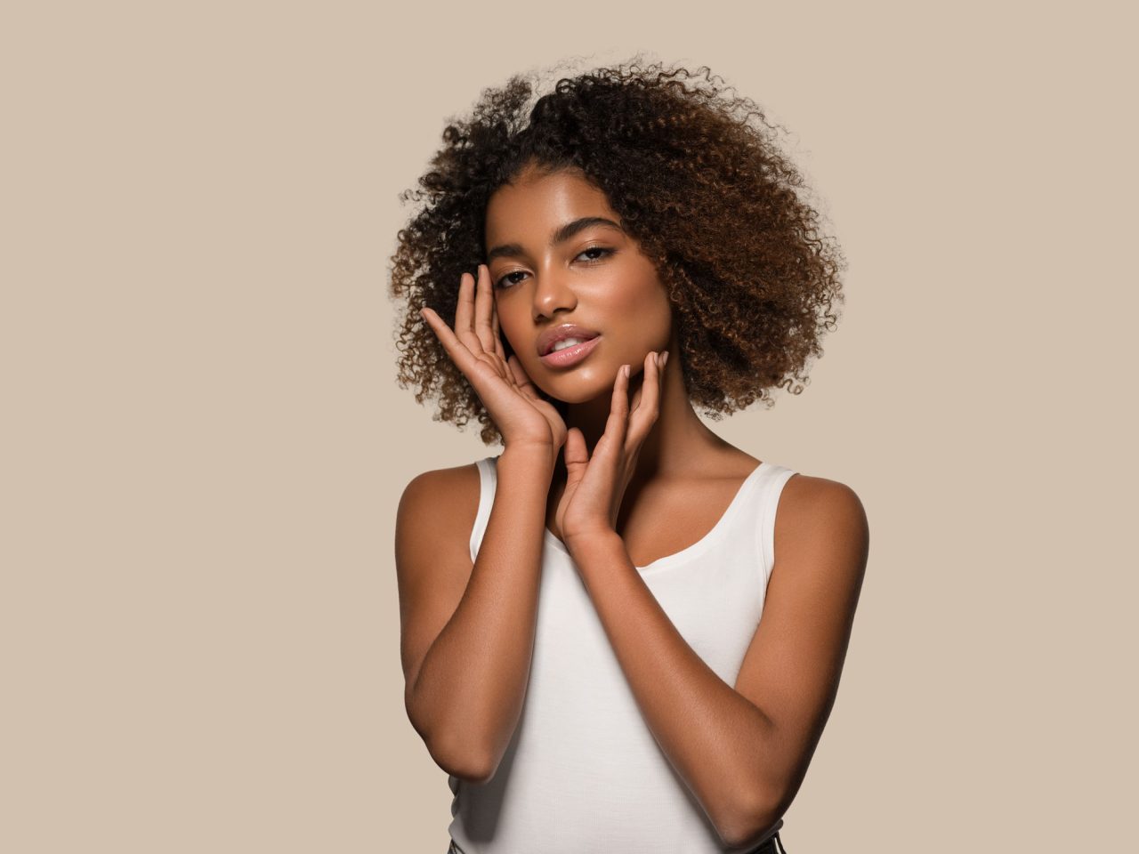 Studio beauty portrait of a woman with curly hair and glowing skin holding her face against a beige background.