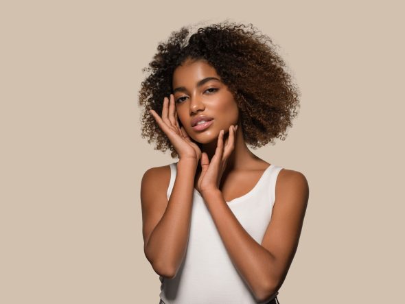 Studio beauty portrait of a woman with curly hair and glowing skin holding her face against a beige background.