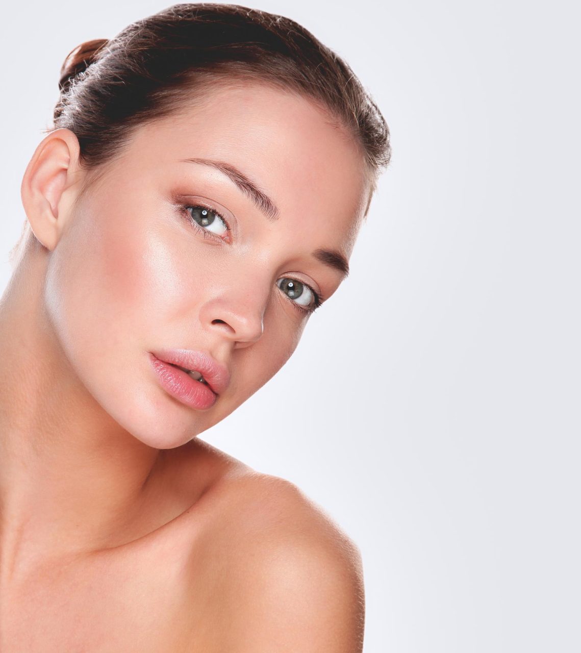 Angled beauty portrait of a woman with glowing skin and hair pulled back against a light gray background