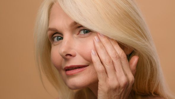Older blonde woman touching her cheek and smiling softly against a beige background