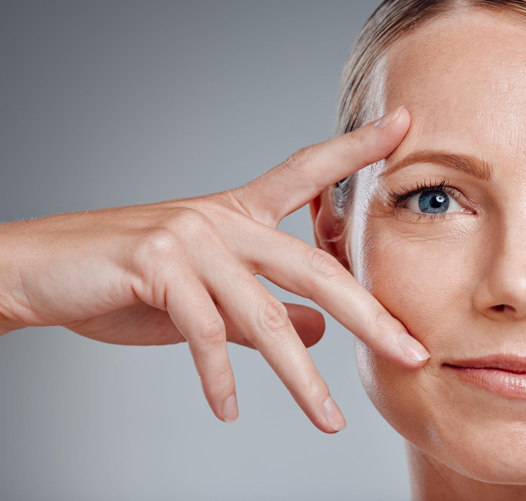 Close-up of an older woman pointing to the skin around her eye showing fine lines against a gray background