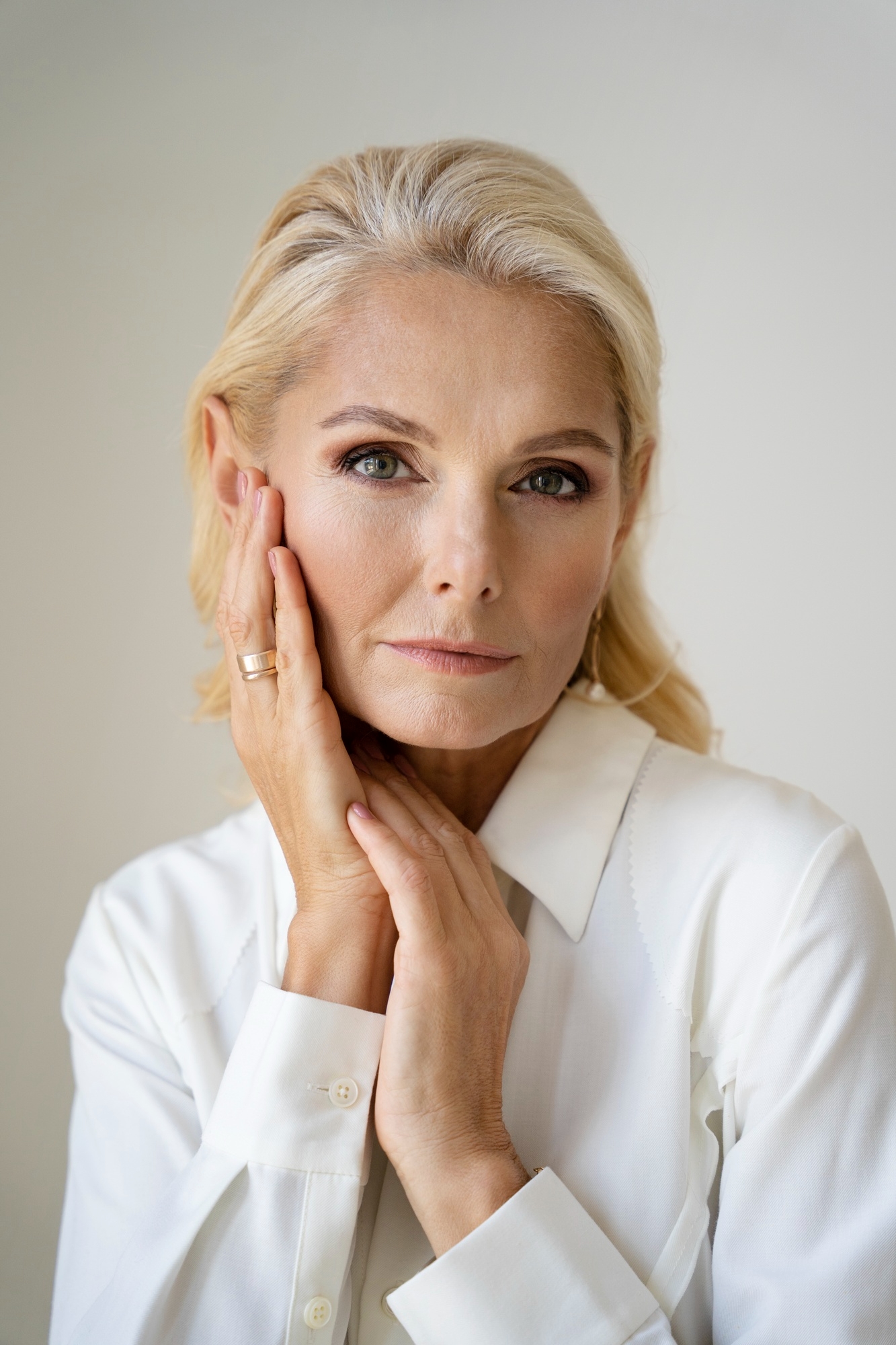 Elegant older blonde woman in a white shirt holding her face with both hands against a light background