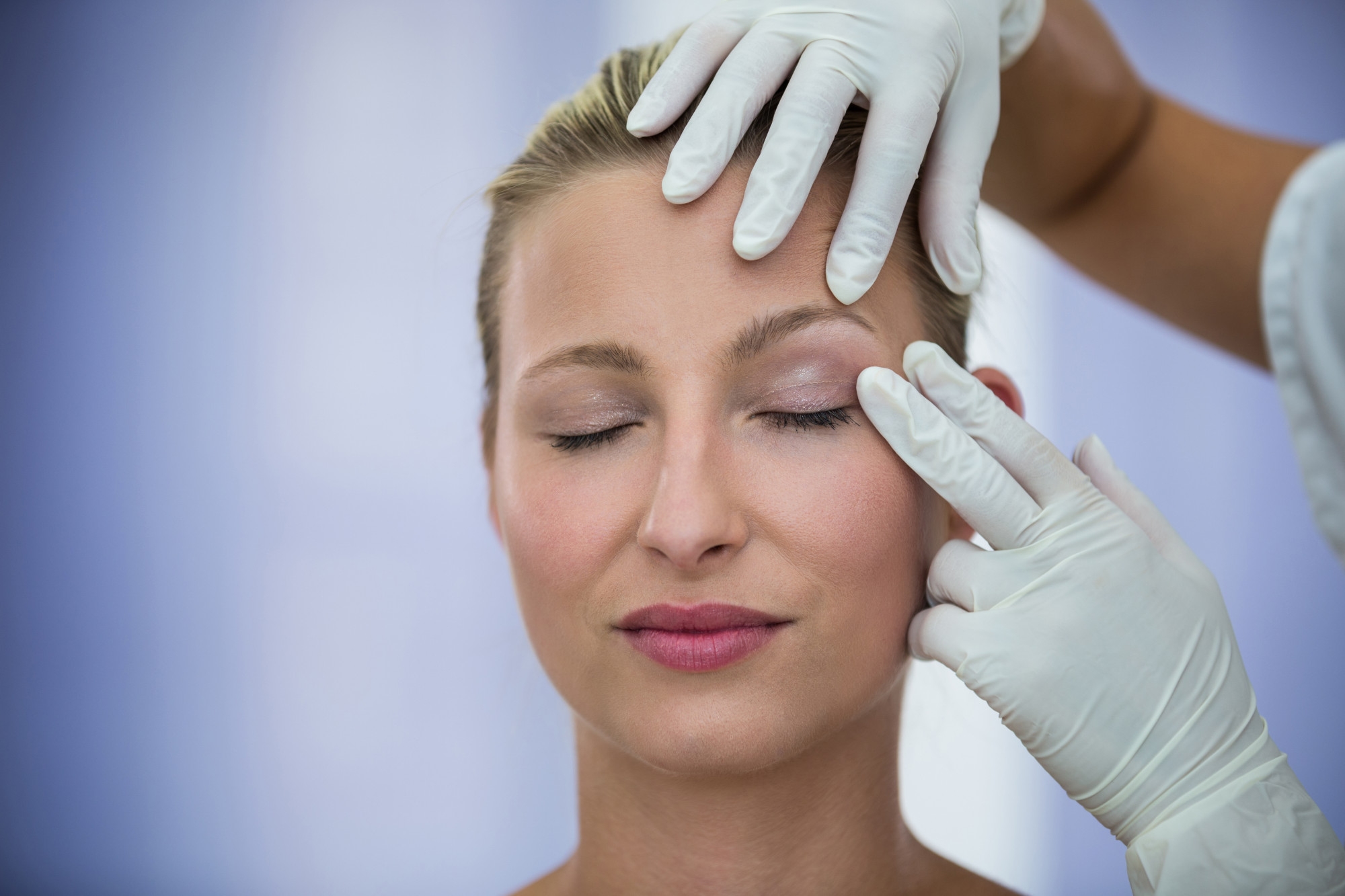 Surgeon in white gloves examining a female patient's eyelid and forehead before blepharoplasty