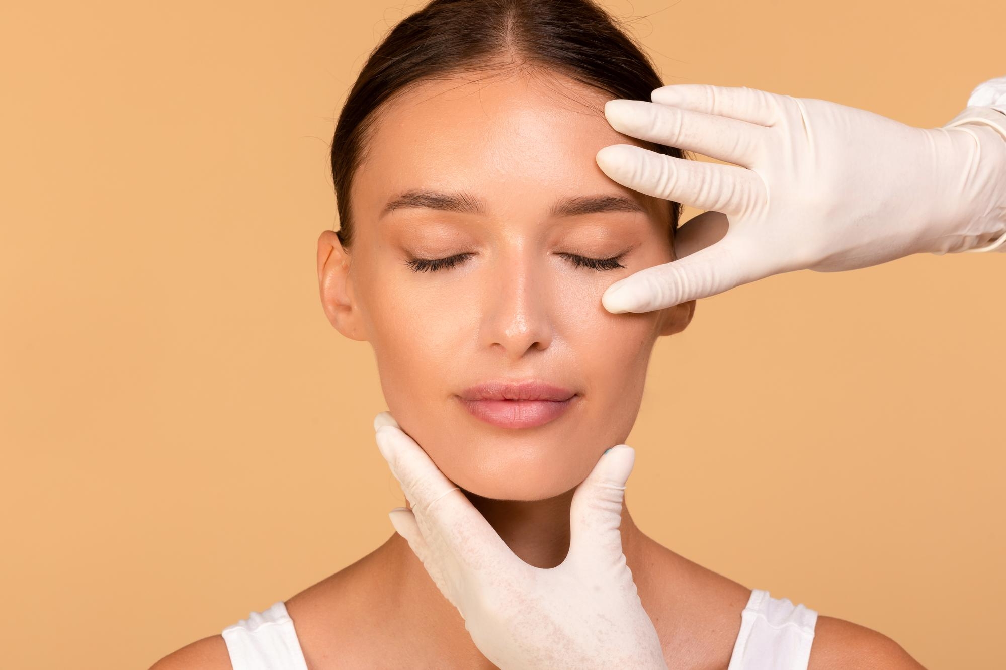 Surgeon in white gloves examining a female patient's eyelid and jaw with eyes closed against a beige background
