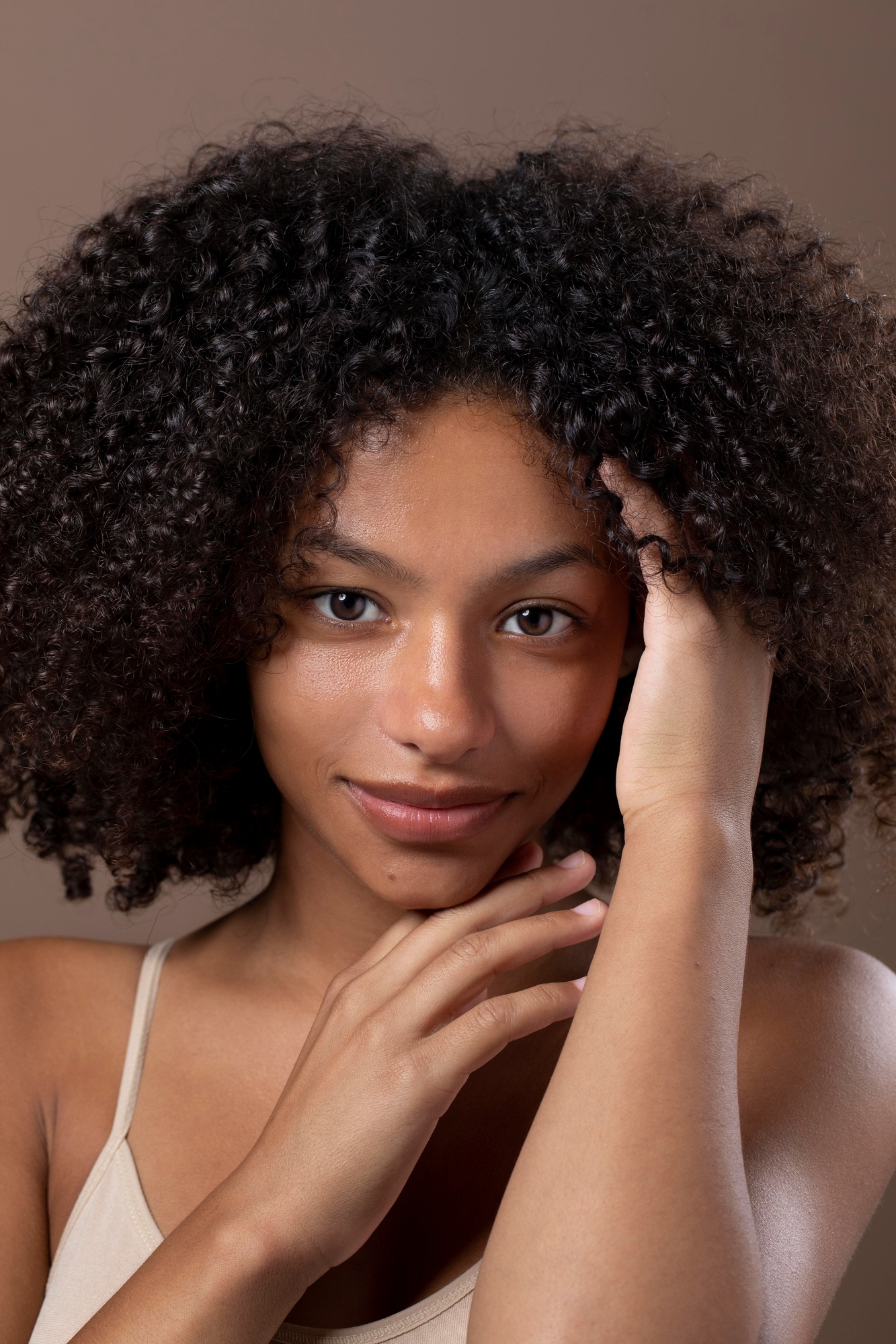 Young woman with natural curly hair and glowing skin touching her face against a brown background