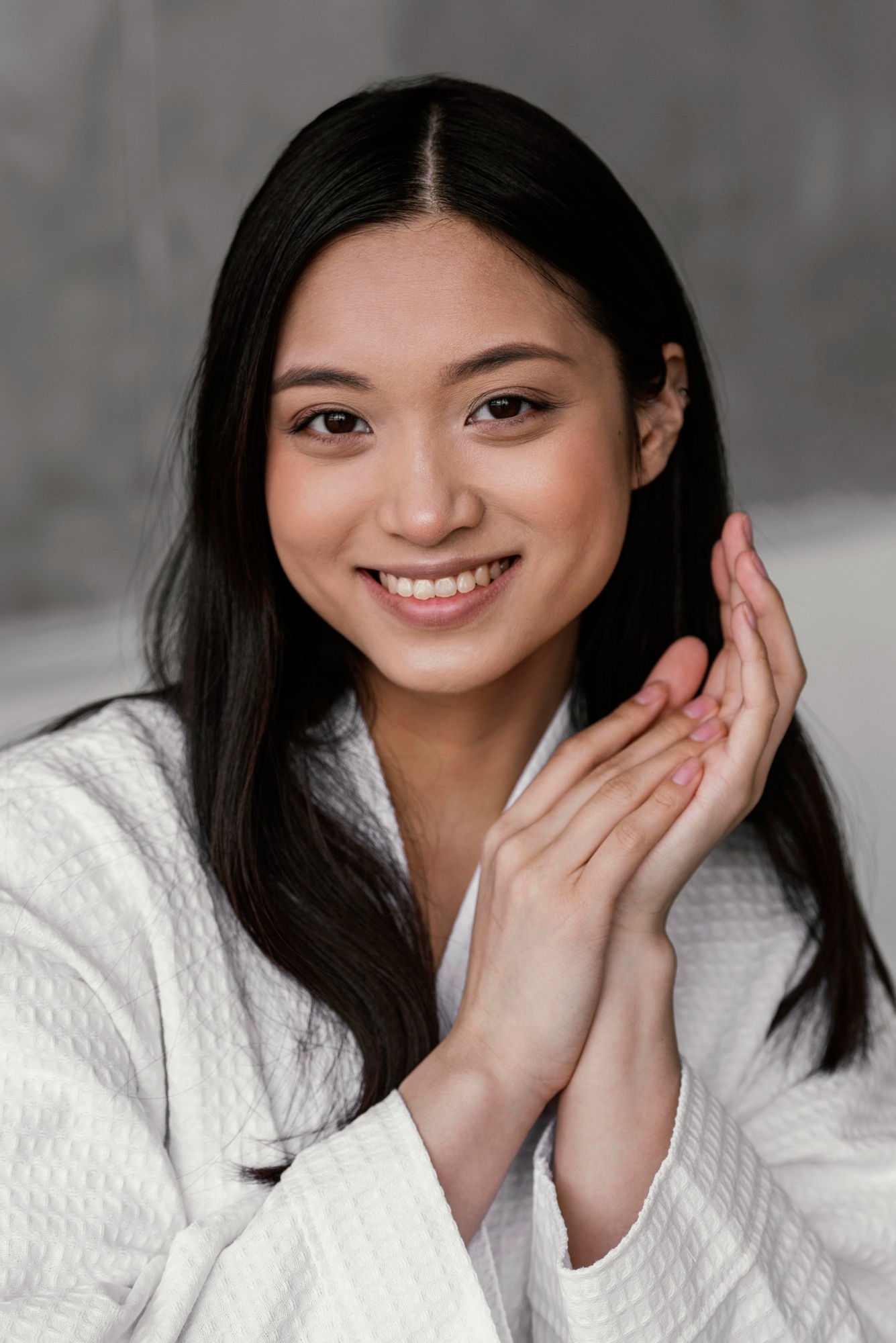 Smiling woman in a white waffle bathrobe with hands together against a gray background