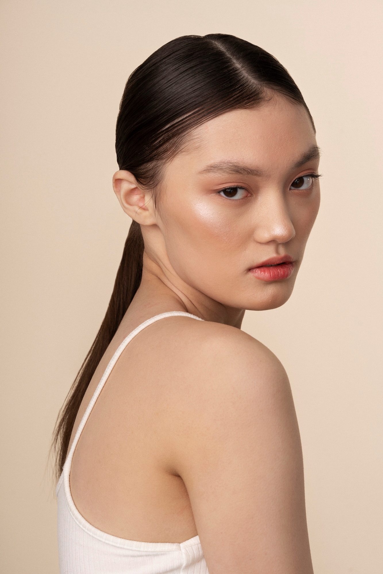 Side profile beauty portrait of an Asian woman with a sleek ponytail and glowing skin looking over her shoulder against a beige background