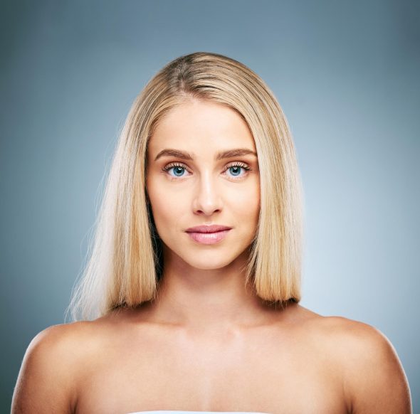 Studio beauty portrait of a blonde woman with a straight bob and clear skin against a blue-grey background