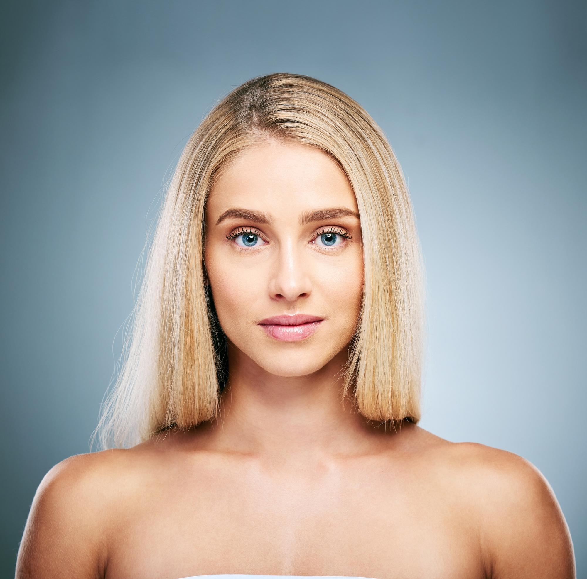 Studio beauty portrait of a blonde woman with a straight bob and clear skin against a blue-grey background