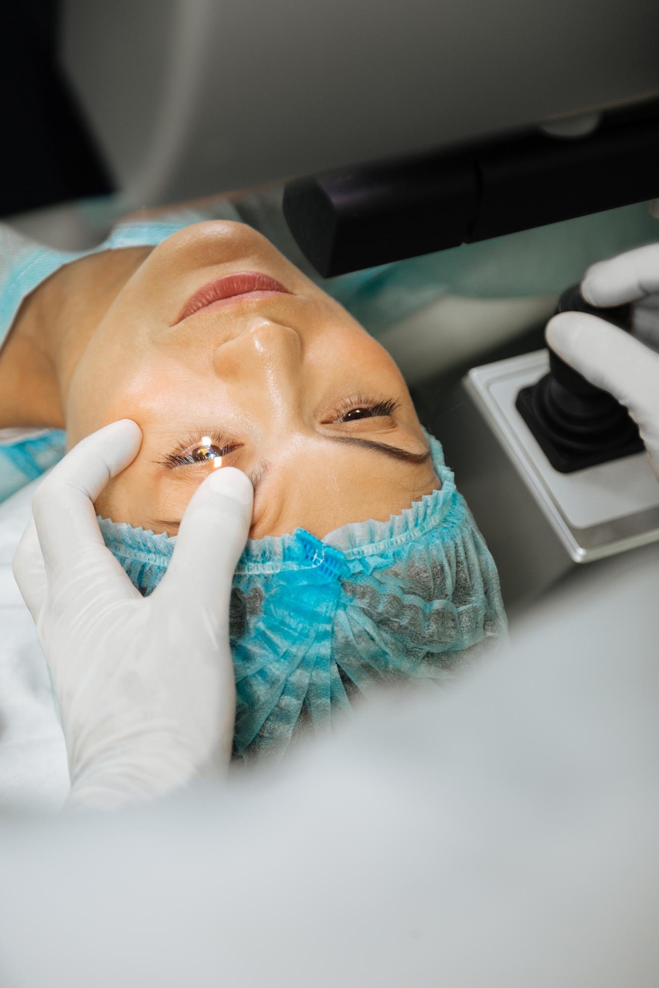 Patient in a surgical cap undergoing laser eye surgery while a gloved practitioner holds the eye open
