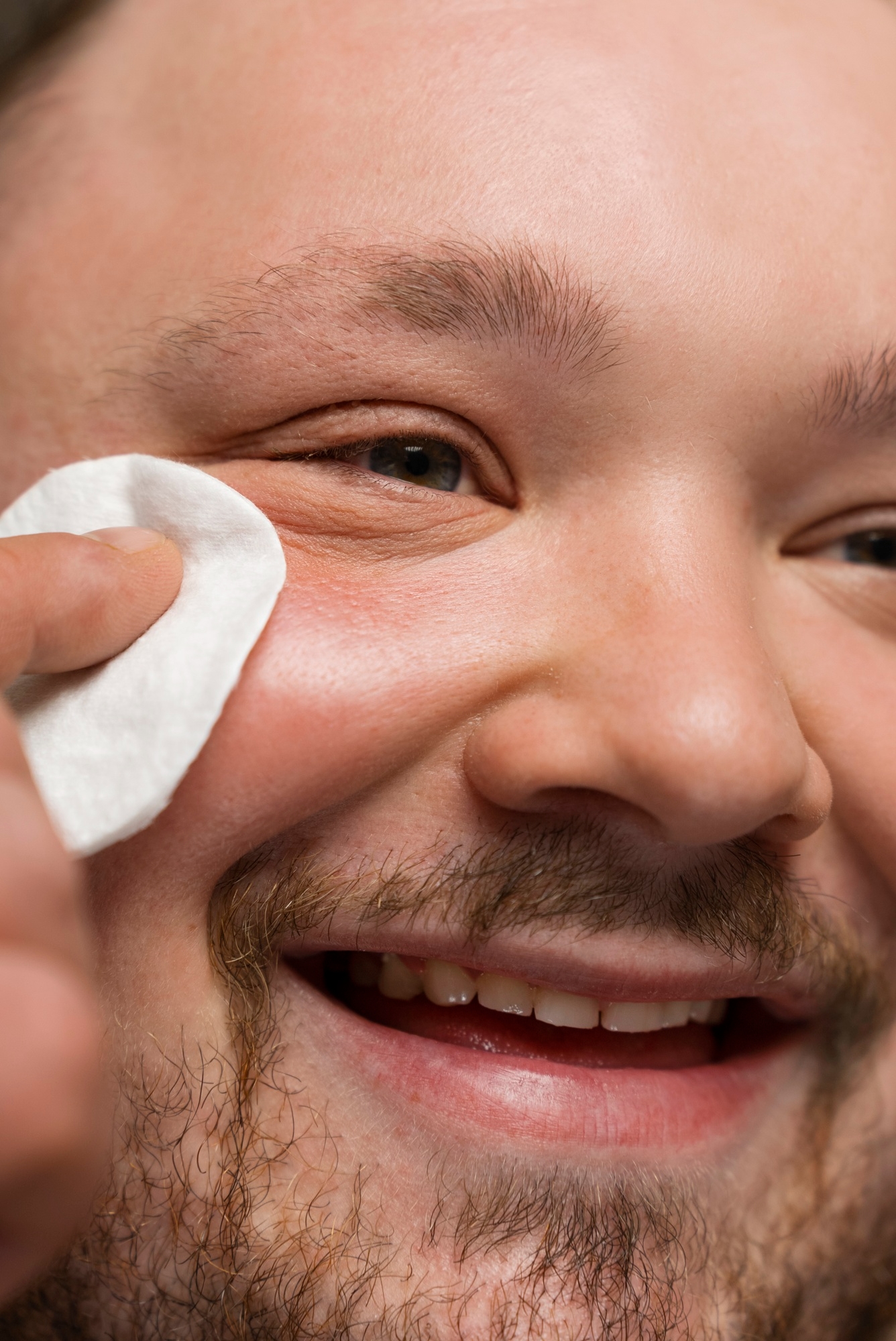 Smiling bearded man wiping under his eye with a white cotton pad in a close-up skincare shot