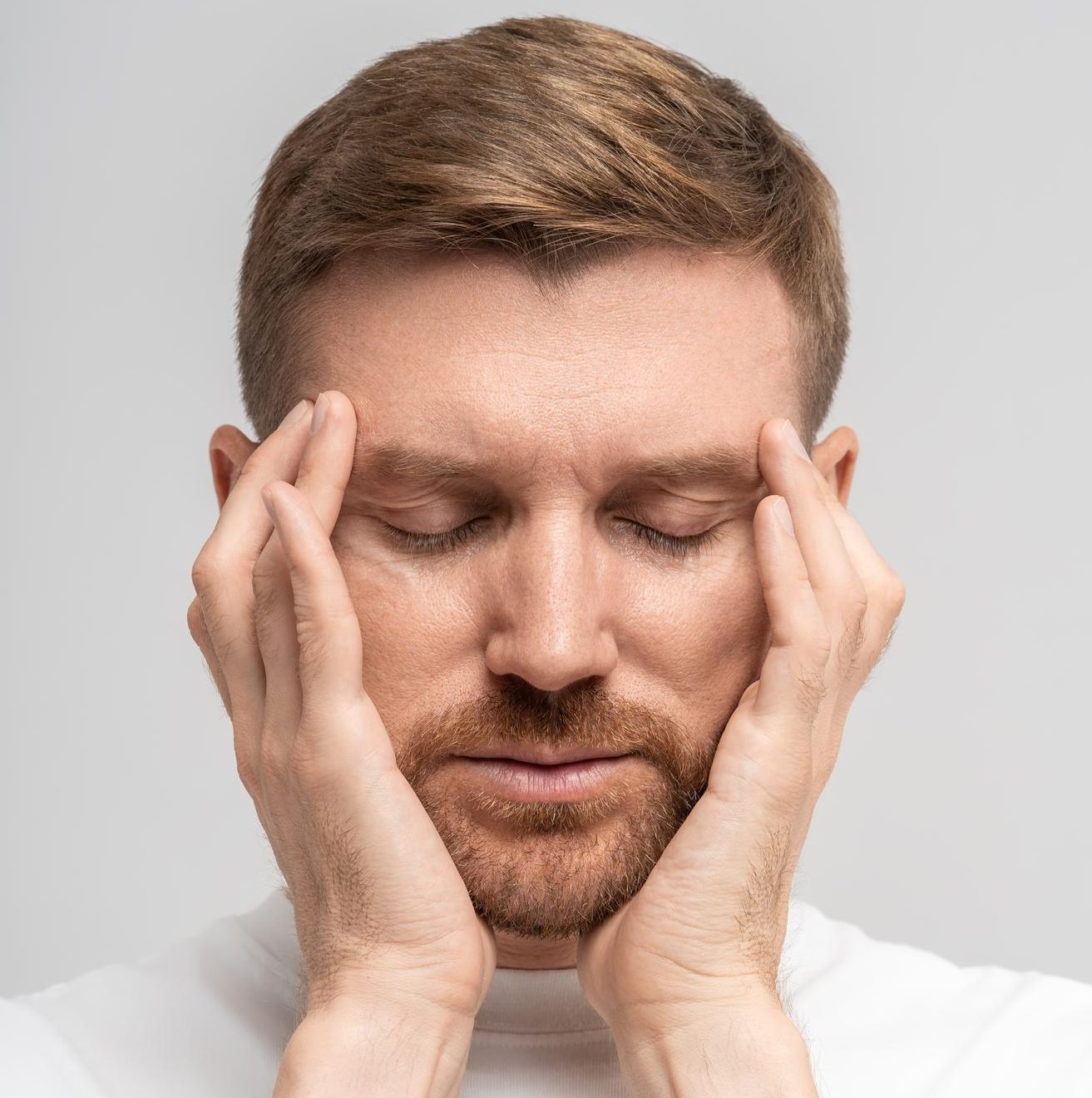 Man with a beard in a white t-shirt pressing his fingertips to his temples with his eyes closed against a grey background