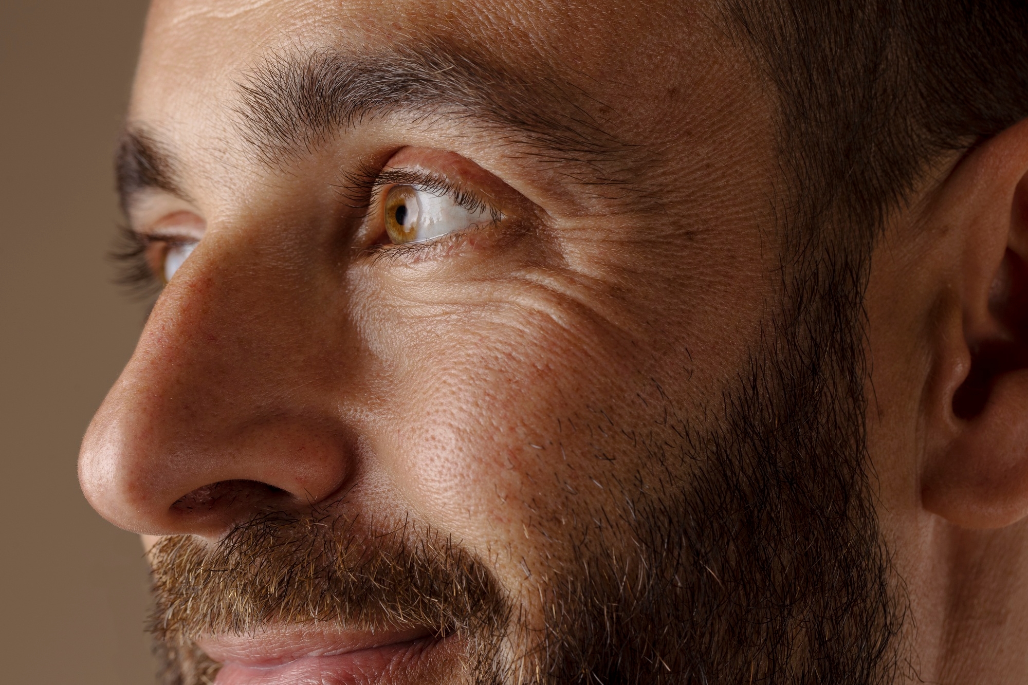 Close-up side profile of a man's hazel eye and face with a beard, looking to the side