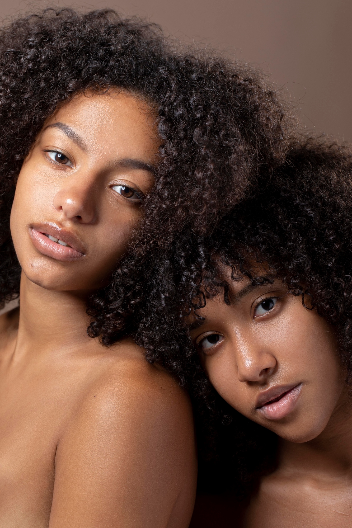 Beauty portrait of two women with curly hair and radiant skin leaning close together against a warm beige background