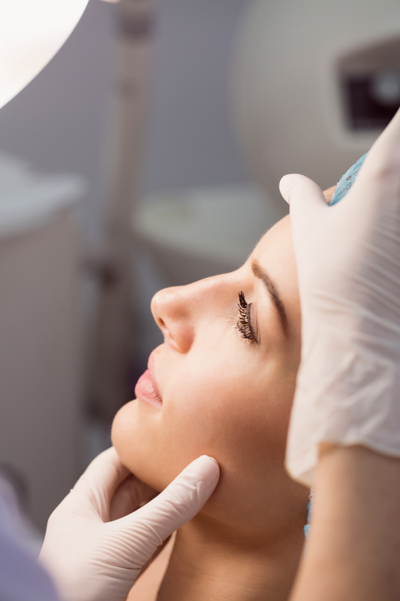 Side profile of a woman with her eyes closed while a gloved practitioner examines her face during an aesthetic consultation