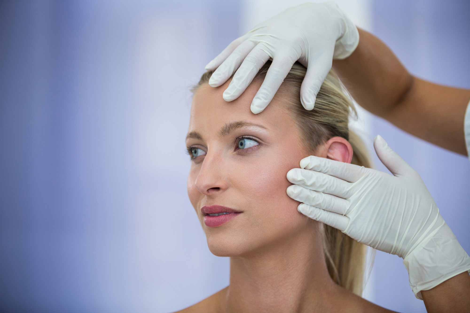 Practitioner in white gloves examining the face and skin of a blonde woman during a cosmetic consultation