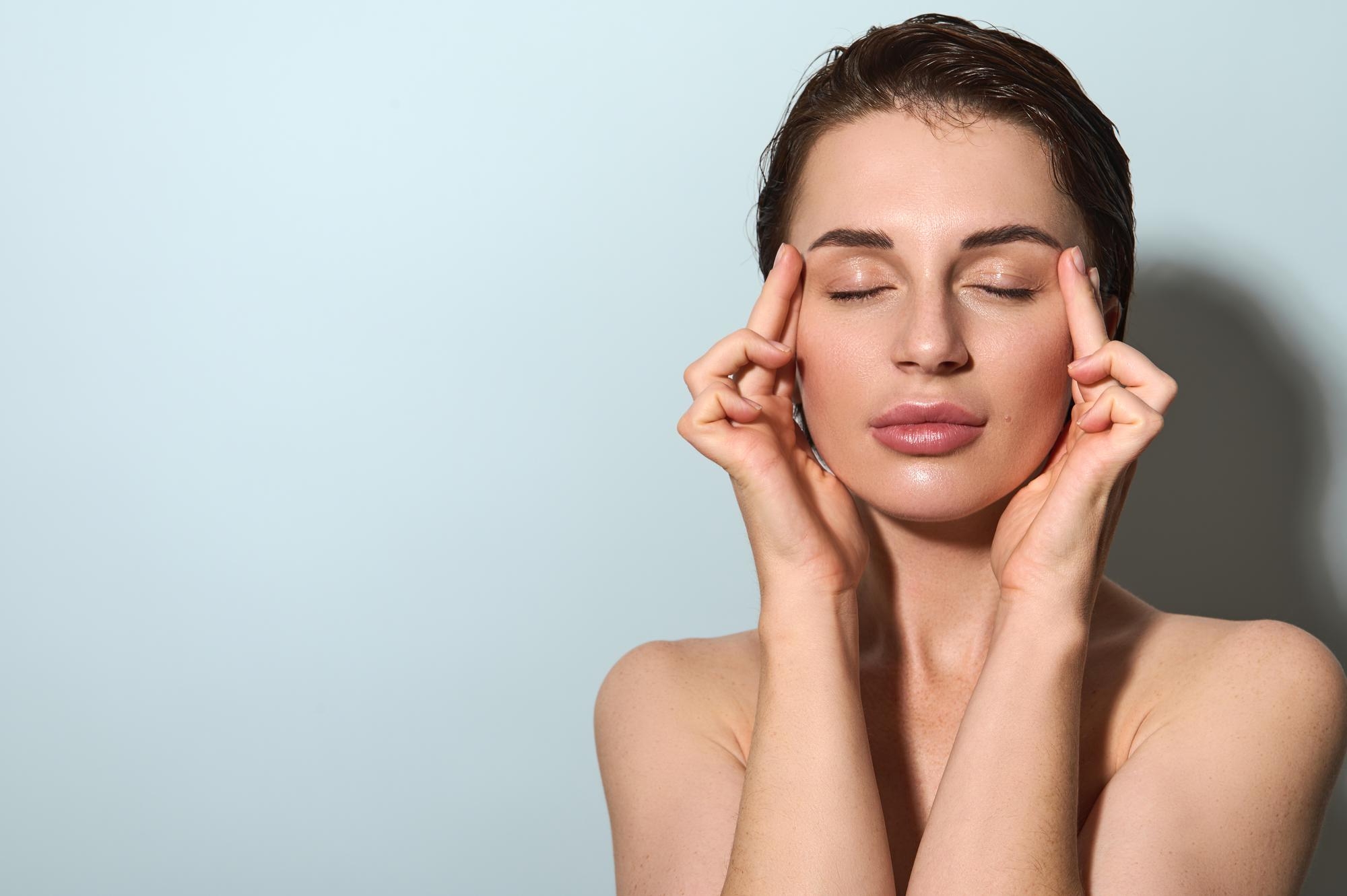 Woman with radiant skin and eyes closed touching her temples with her fingertips against a pale blue background