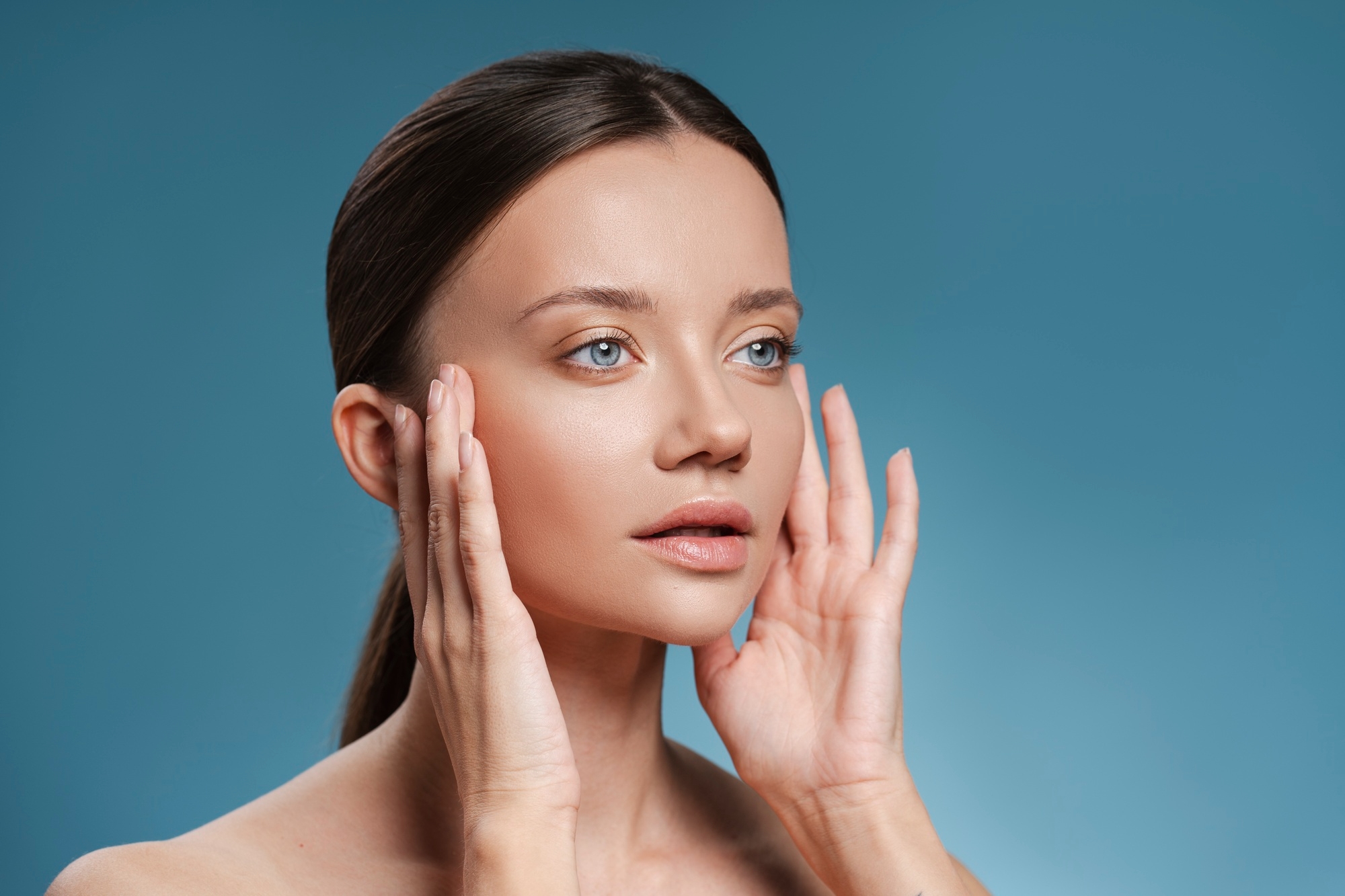 Beauty portrait of a brunette woman with glowing clear skin framing her face with her hands against a blue background