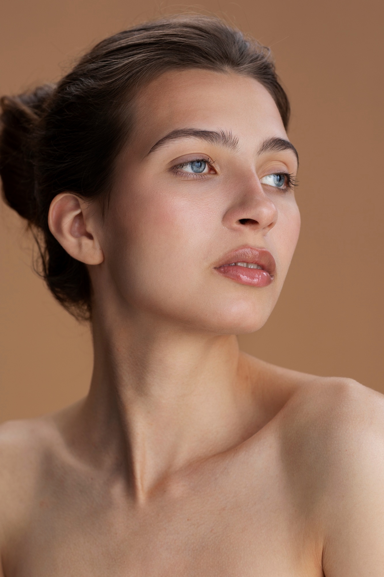 Beauty portrait of a woman with her hair in a bun and dewy natural skin looking upward against a warm beige background