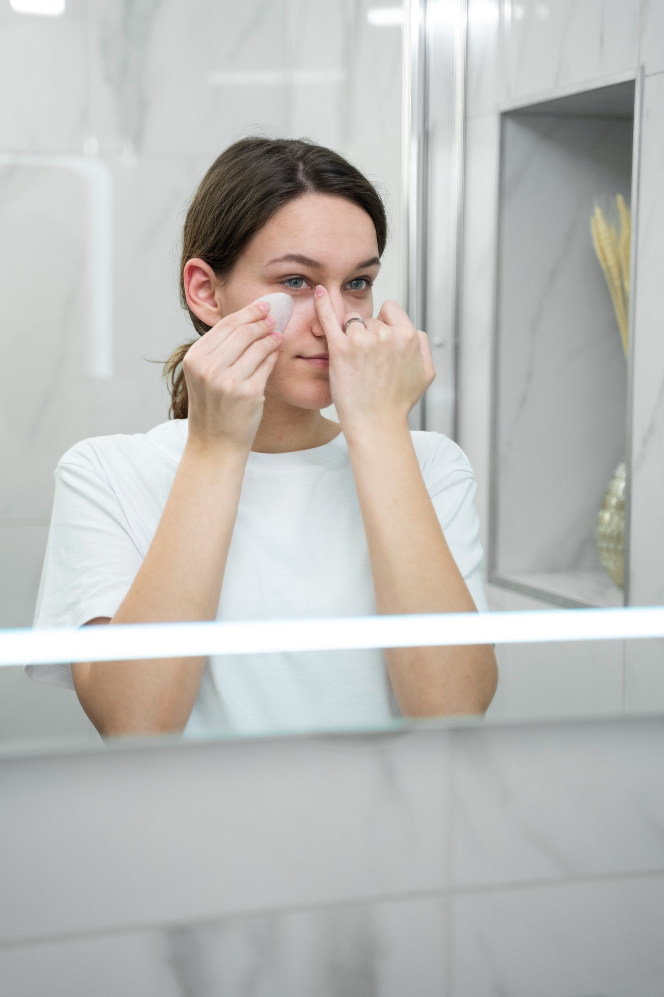 Woman in a white t-shirt using a gua sha tool under her eye while looking in a bathroom mirror
